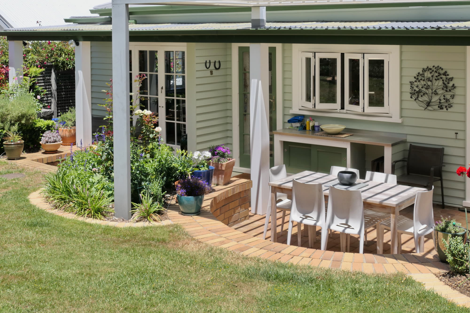 The garden elevation of the villa: pale green weatherboard, a white pergola, french doors and a servery window above an outdoor dining table.