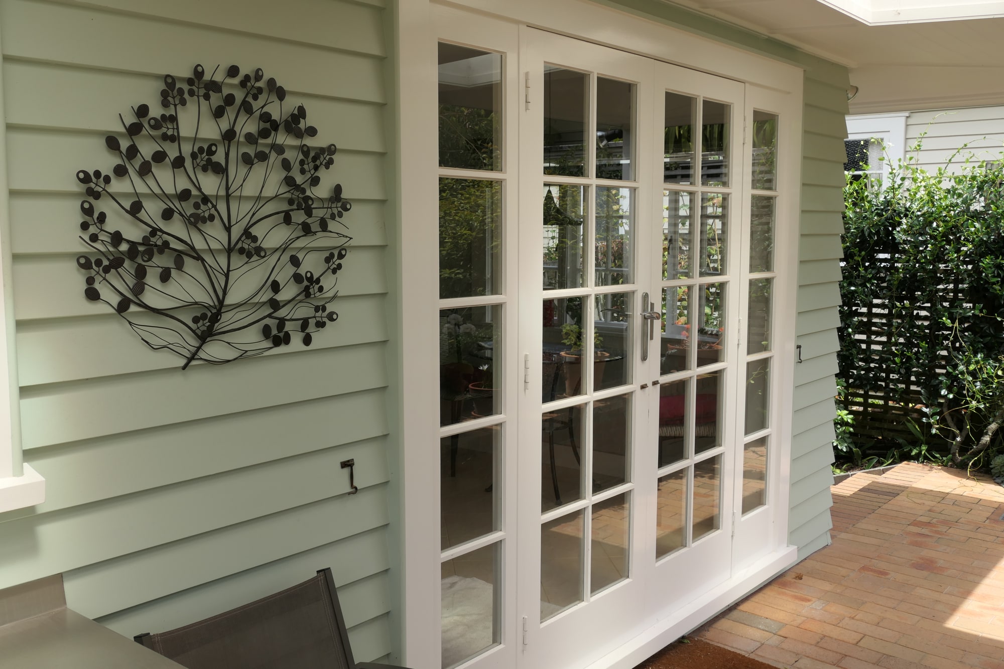 White french doors in a pale green weatherboard wall, with a metal wall sculpture alongside.