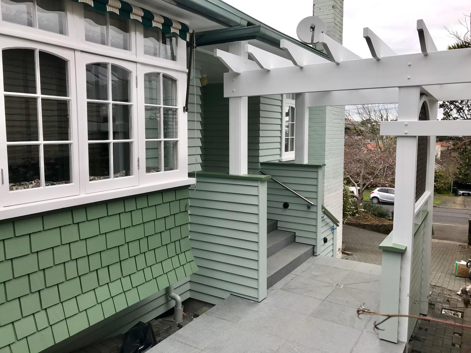 A white pergola over granite steps against pale green weatherboard, shingles and white bay-window joinery.