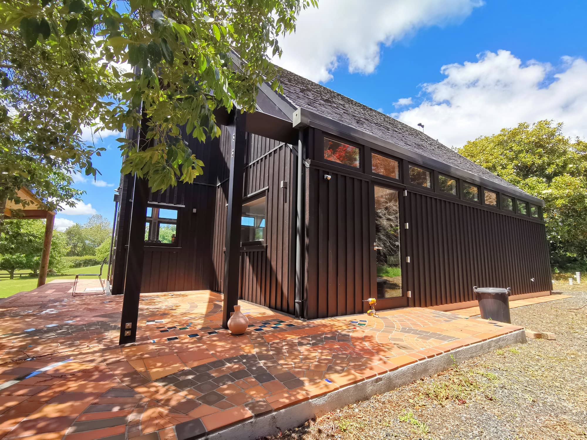 The long elevation of a dark brown stained timber studio under dappled light from a tree.