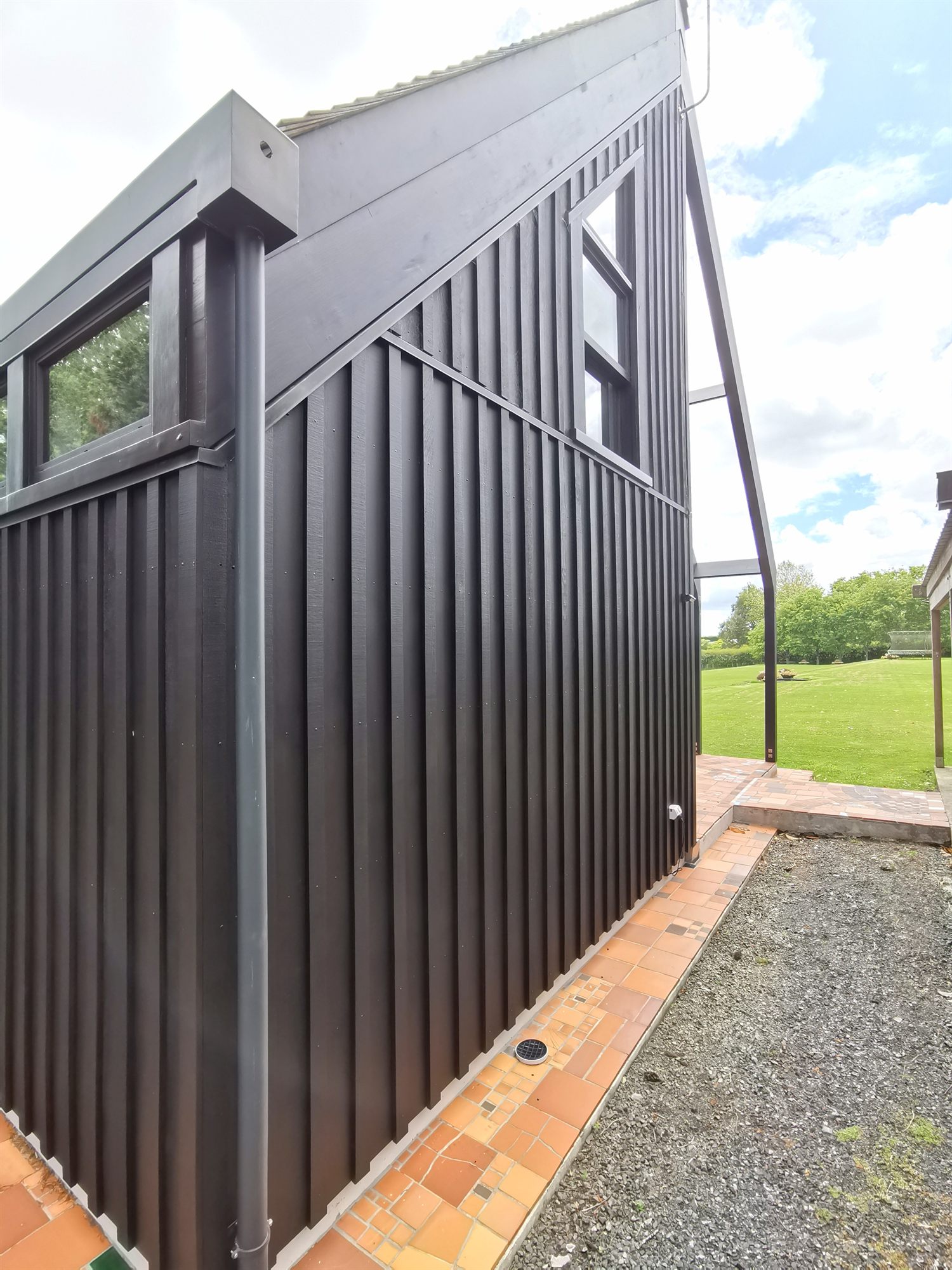 The angled dark brown board-and-batten gable of the studio above a tiled apron, looking out across the lawn.