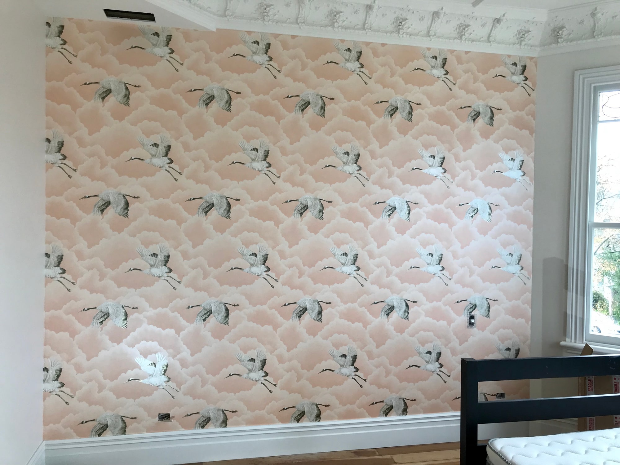 Pale pink wallpaper patterned with flying cranes across a bedroom feature wall.