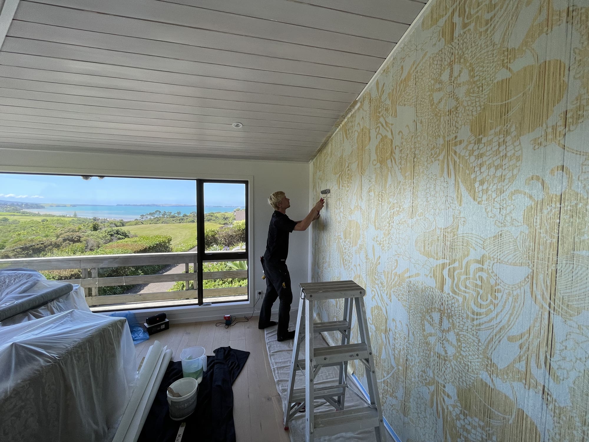 A decorator smoothing a length of gold botanical wallpaper onto a wall beside a window with a sea view.